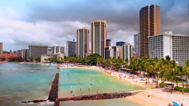 Pasifik Okyanusu kıyısındaki güzel bir tatil köyü. Turkuaz suda yüzen ve kumsalda güneşlenen çok sayıda turist var. Göz kamaştırıcı binalar arka planda duruyor. Waikiki, Hawaii, ABD.