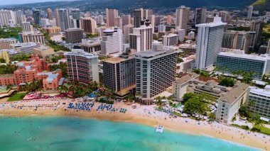Tatilcilerin ve mavi şemsiyelerin olduğu uzun, dar kumlu bir sahil. Waikiki, Hawaii, ABD 'nin çeşitli modern binaları. Hava görünümü.