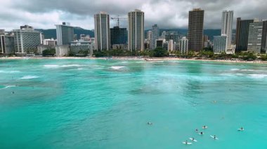 Sörfçüler sörf tahtalarında oturup yaklaşan dalgayı beklerler. Hava aracı görüntüleri Waikiki, Honolulu, Hawaii 'de bulutlu gökyüzünün altında sahile yaklaşıyor..