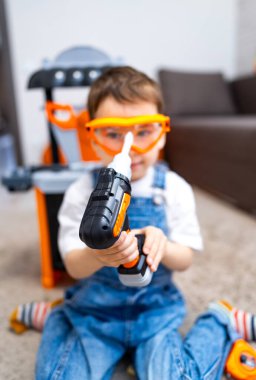 Çocuk oyuncak matkapla oynuyor. Güvenlik gözlüğü takan küçük bir çocuk, yumuşak mobilyalarla dolu, kapalı bir alanda oyuncak matkapla oynuyor..