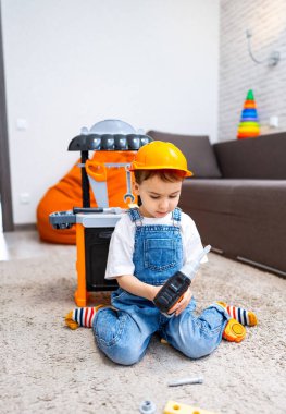 Çocuk içeride oyuncak aletlerle oynuyor. Küçük çocuk halının üzerinde oturuyor, oyuncak matkabına odaklanmış haldeyken kapalı bir alanda renkli oyun ekipmanlarıyla çevrili..
