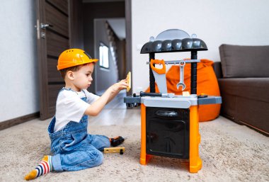 Çocuk içeride oyuncak aletlerle oynuyor. İnşaat şapkalı çocuk oturma odasında halıda otururken oyuncak aletleri yaratıcı bir şekilde kullanıyor..