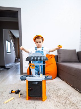 Çocuk içeride yaratıcı bir şekilde oynuyor. Sert şapkalı genç bir çocuk, parlak bir oturma odasında oyuncak bir aletle oynuyor, yaratıcı inşaatın keyfini çıkarıyor..