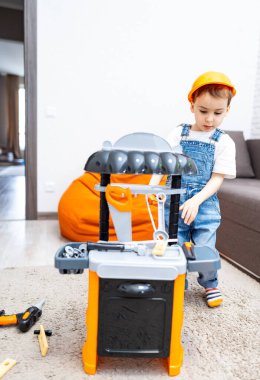 Çocuk içeride oyuncak setiyle oynuyor. Tulumlu genç bir çocuk, rahat bir oturma odasında oyuncak inşaatı ile meşgul..