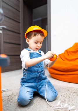 Çocuk oyuncaklarla oynuyor. İnşaat şapkalı genç bir çocuk, kapalı bir alanda halının üzerinde otururken aletlerle oynuyor..