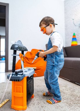 Çocuk içeride oyuncak aletlerle oynuyor. Tulum ve güvenlik gözlüğü takan genç bir çocuk evinde rahat bir oturma odasında oyuncak matkapla eğleniyor..