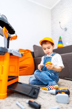 Çocuk içeride oyuncak aletlerle oynuyor. Turuncu şapkalı genç bir çocuk, kapalı bir alanda halının üzerinde oyuncak aletler toplamaktan hoşlanır..