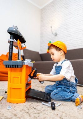 Çocuk içeride oyuncak aletlerle oynuyor. Tulum giymiş ve şapka takmış bir çocuk, içerideki bir halıda otururken oyuncak bir aletle meşgul oluyor..