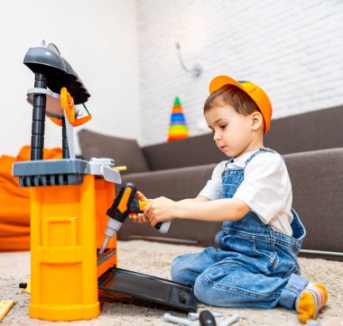 Çocuk oyuncak aletlerle oynuyor. Oyun odasındaki genç çocuk inşaat şapkası ve kot pantolon giyerken tezgahında oyuncak matkap kullanıyor..