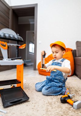 Çocuk oyuncak ızgarayla eğleniyor. Tulumlu ve şapkalı bir çocuk evin içinde renkli mobilyalarla çevrili bir oyuncak ızgara yapmaya odaklanır..