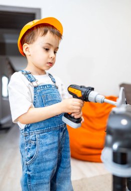 Küçük bir çocuk oyun alanında matkap kullanıyor. Genç bir çocuk, parlak bir iç mekanda oyuncak matkap kullanırken inşaat şapkası ve tulumu takıyor..