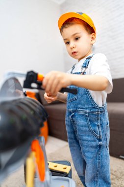 Çocuk alet takımıyla oynuyor. Tulum giymiş ve kasketli genç bir çocuk içeride oyuncak bir aletle oynuyor, bir inşaat projesine odaklanmış..