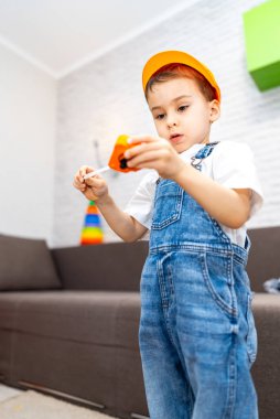 Çocuk içeride oyuncak arabayla oynuyor. Mavi tulumlu ve turuncu şapkalı genç çocuk parlak bir oturma odasında oynarken oyuncak arabasına odaklanıyor..