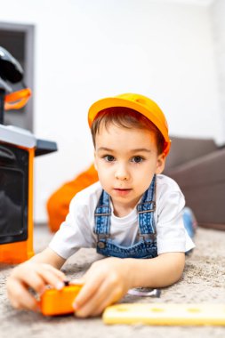 Çocuk oyuncak aletlerle oynuyor. İnşaat şapkalı genç çocuk neşeli bir oyun odasında yumuşak bir halıda oyuncak aletlerle meşgul oluyor..