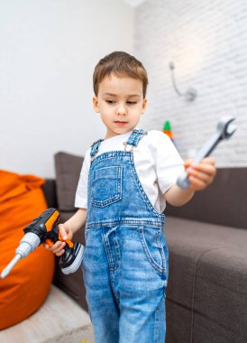 Küçük çocuk aletlerle oyun oynamaktan hoşlanıyor. Kot pantolon giymiş genç bir çocuk, parlak bir oturma odasında yaratıcı bir oyun oynayan bir alet ve İngiliz anahtarı tutuyor..