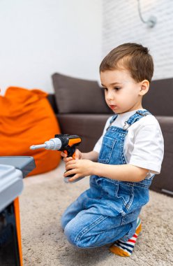 Çocuk evde yaratıcı bir şekilde oynuyor. Kot pantolon giymiş bir çocuk oyuncak matkap kullanır, kapalı oyun saatinde bir çalışma tezgahına dikkatle odaklanır.
