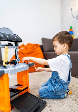 Çocuk içeride oyuncak ızgarasıyla oynuyor. Küçük bir çocuk halının üzerinde diz çöküp, oyuncak ızgara yapıyor ve yemek pişirme hareketlerini taklit ediyor..