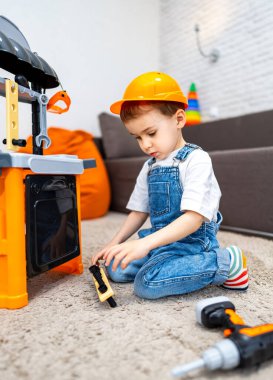 Çocuk içeride oyuncak aletlerle oynuyor. İnşaat şapkalı genç çocuk evde oyuncak aletlerle oynamaktan hoşlanıyor, oyundaki yaratıcılığı ve hayal gücünü vurguluyor.