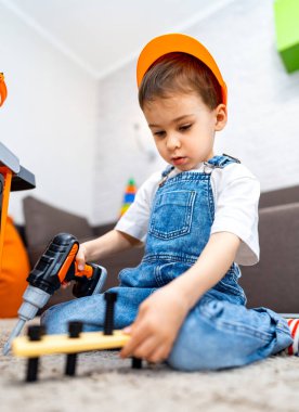 Çocuk oyuncak aletlerle oynuyor. Parlak şapkalı genç bir çocuk evdeki bir inşaat projesinde halıda otururken oyuncak matkap kullanıyor..
