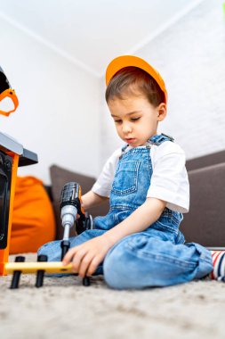 Çocuk içeride oyuncak aletlerle oynuyor. Küçük çocuk yerde otururken oyuncak matkap kullanmaya odaklanmış. Üzerinde güvenlik şapkası ve tulum var..