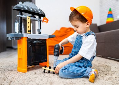 Çocuk DIY aktiviteleri oynuyor. Sert şapkalı genç bir çocuk mutfak setinde çalışmak için oyuncak matkap kullanıyor, içeride yaratıcı oyunlardan hoşlanıyor..