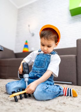 Samimi odada matkapla gezen çocuk. Genç bir çocuk, renkli dekorlarla dolu modern bir yaşam alanında matkapla oyuncak parçaları birleştirmeye odaklanır..