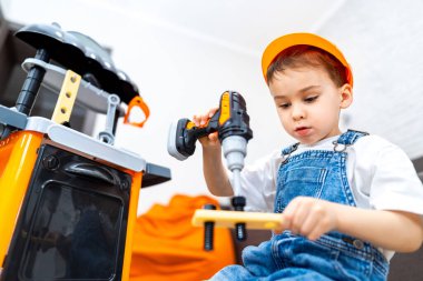 Çocuk içeride oyuncak aletlerle oynuyor. İnşaat şapkalı küçük bir çocuk tezgahın yanında oynarken tahta bir parça üzerinde oyuncak matkap kullanıyor..