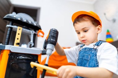 Çocuk oyuncaklarla mutlu bir şekilde oynuyor. Sert şapka takan bir çocuk evde oyuncak matkap ve aletler kullanmaya odaklanır..