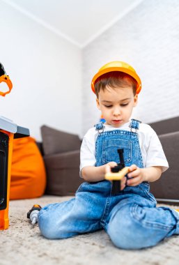 Çocuk oyuncak bloklarıyla oynuyor. İnşaat şapkalı küçük bir çocuk, rahat bir oturma odasında yumuşak bir halıya serilmiş bir oyuncakla oynuyor..