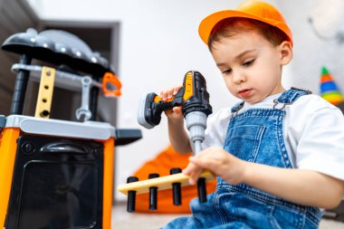 Çocuk aletlerle yaratıcı bir şekilde oynuyor. Sert şapkalı genç bir çocuk, oyuncak matkap kullanarak oyun odasında oyuncaklar üzerinde çalışıyor. Yaratıcı inşaatın keyfini çıkarıyor..