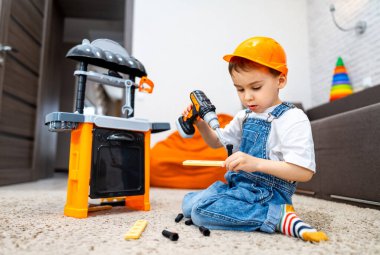 Çocuk oyuncaklarla yaratıcı bir şekilde oynuyor. Turuncu şapkalı küçük çocuk oyun atölyesinin yanındaki halıda otururken oyuncak matkap kullanıyor..
