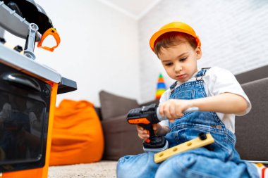 Çocuk matkapla yaratıcı bir şekilde oynuyor. Kot pantolonlu ve şapkalı bir çocuk kapalı bir alanda oynarken oyuncak matkap kullanmaya odaklanır..