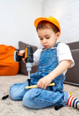 Aletleri oyunda olan genç bir inşaatçı. Tulumlu bir çocuk yerde otururken oyuncak matkap kullanıyor, içeride ahşap işçiliği faaliyetlerine odaklanıyor..