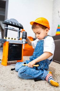 Küçük çocuk yaratıcı bir oyun oynar. İnşaat kaskı takmış bir çocuk halının üzerinde otururken oyuncak matkap kullanıyor. Yaratıcılık ve odak gösteriyor..