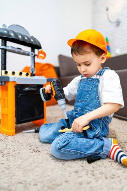 Çocuk oyuncak aletlerle oynuyor. Tulumlu ve şapkalı bir çocuk, yumuşak halıda otururken oyuncak matkap kullanıyor gibi yapıyor..
