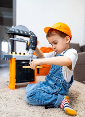 Genç inşaatçı yaratıcı oyun zamanlarını seviyor. Sert şapkalı bir çocuk tezgah setinde oyuncak matkap kullanır, yaratıcılığını gösterir ve kapalı alanda yaratıcı oyunlar oynar..