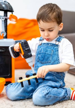 Çocuk yaratıcı oyunlardan hoşlanıyor. Kot pantolon giymiş küçük bir çocuk, kapalı bir oyun alanında yumuşak bir halıda otururken parçaları takmak için oyuncak matkap kullanıyor..