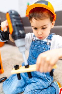 Oyuncak matkap kullanan genç bir inşaatçı. Tulumlu ve şapkalı bir çocuk tahta bir projede oyuncak matkap kullanıyor, odaklanmış ve oyun oynuyor gibi yapıyor..