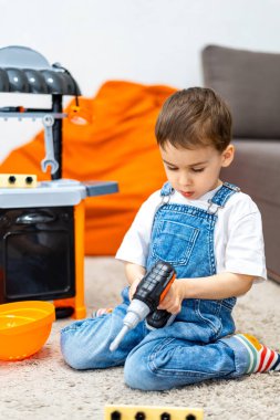 Çocuk içeride oyuncak matkapla oynuyor. Tulumlu bir çocuk halıda oyun mutfağı olan bir yerde otururken oyuncak matkap kullanıyor..