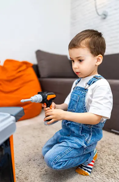 Çocuk evde yaratıcı bir şekilde oynuyor. Kot pantolon giymiş bir çocuk oyuncak matkap kullanır, kapalı oyun saatinde bir çalışma tezgahına dikkatle odaklanır.