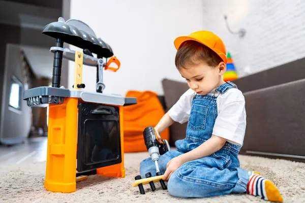 Çocuk yapı taşlarıyla oynuyor. Parlak şapkalı bir çocuk evde renkli bir tezgahın yanında oynarken oyuncak matkap kullanıyor..