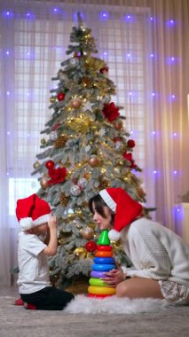 Sevimli küçük çocuk gözlerini Noel Baba şapkasının altına gizliyor. Anne ve çocuk Noel ağacının yanında saklambaç oynuyorlar. Dikey video.