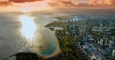 Pasifik Okyanusu dalgaları Waikiki, Honolulu, Hawaii, ABD kıyılarına ulaşır. Batan güneş manzarayı aydınlatıyor. Dağların üstündeki gökyüzünde turuncu bulutlar.