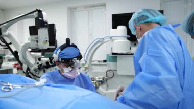 Hastaya bakan cerrah cerrahi lensler takıyor. Mavi üniforma giyen erkek asistan doktora yardım ediyor..