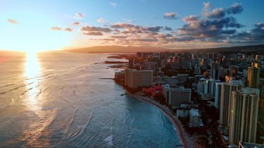 Alacakaranlıkta Waikiki bölgesinin üzerindeki görüntüler. Batan güneş, Pasifik Okyanusu 'nun sularını aydınlatıyor. Pofuduk bulutlar şehrin üzerindeki gökyüzünü kaplıyor. Üst görünüm.