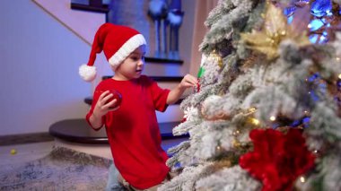 Uzun kırmızı tişört giyen ve Noel Baba şapkası takan sevimli çocuk Noel ağacında oturur. Bebek ağaçtan birkaç oyuncak alıyor..