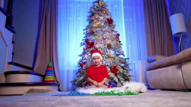 Kırmızı şapkalı ve tişörtlü gülen çocuk Noel ağacının yanında oturuyor. Güzel bebek boncuk dizisiyle oynuyor. Düşük açı görünümü.