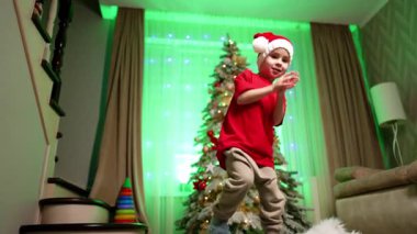 Kırmızı tişörtlü ve Noel Baba şapkalı şirin çocuk el çırpıyor ve mutlu bir şekilde dans ediyor. Düşük açı Noel 'de evde eğleniyor..