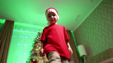Kırmızı şapkalı ve tişörtlü mutlu bebek içeride neşeyle zıplıyor. Kameranın önünde dans eden neşeli çocuk. Düşük açı görünümü.