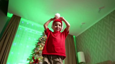 Beyaz çocuk yüzünü Noel Baba şapkasının arkasına gizliyor. Evde zıplayan çocuğa alçak açılı bir bakış açısı. Arkaplanda Noel ağacı.